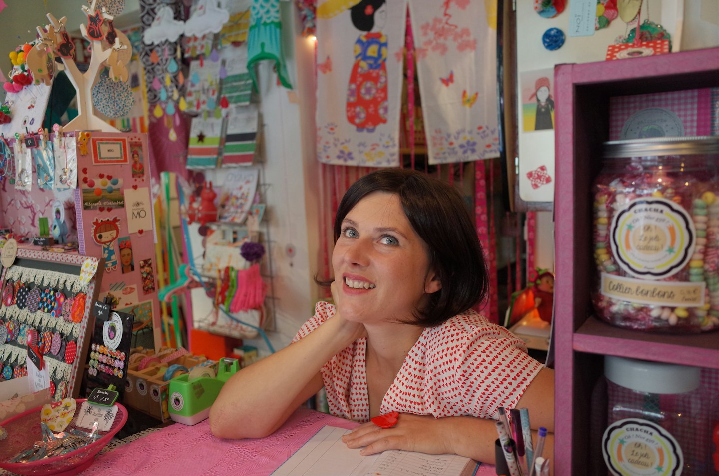 Mujer sonriente apoyada en un mostrador rosa en una concurrida tienda de regalos.